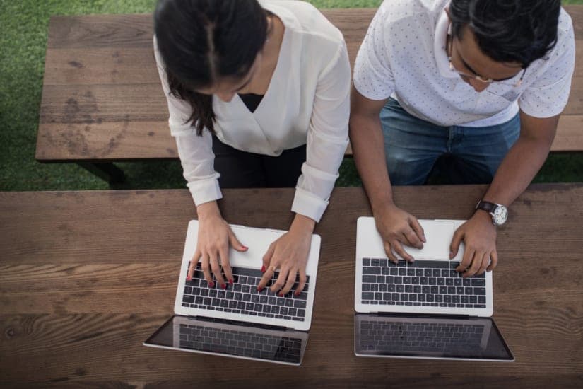 Two people working at laptops on a wooden table, photographed from directly above so neither face is visible. A stand-in, not this office.