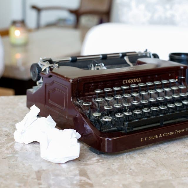 A dark red vintage Corona typewriter on a pale stone counter, with a crumpled sheet of paper beside it. A stand-in, not this office.