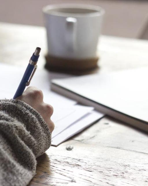 A hand in a knitted sleeve writing in a notebook on a wooden table, next to a mug of coffee. A stand-in, not this office.