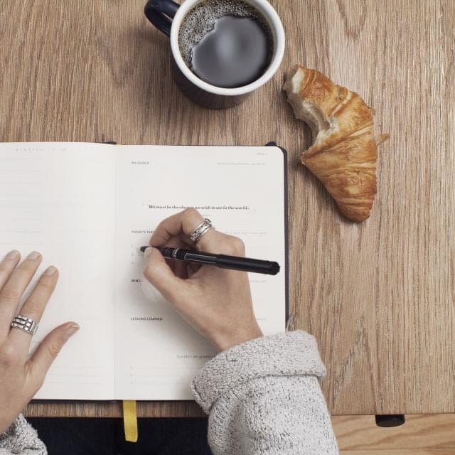 Seen from above, a hand writing in an open notebook on an oak table, beside a mug of black coffee and a half-eaten croissant. A stand-in, not this office.